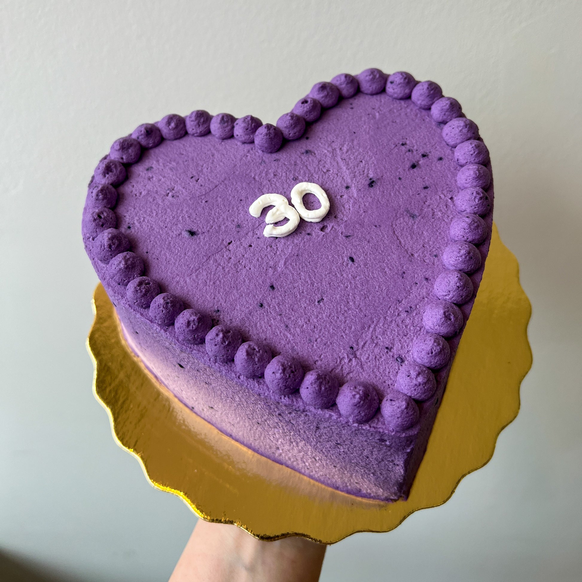 A heart-shaped Ube crepe cake with piped frosting borders and a white '30' decoration, displayed on a gold scalloped cake board held by a hand against a plain wall.