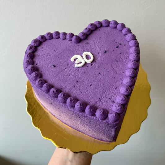 A heart-shaped Ube crepe cake with piped frosting borders and a white '30' decoration, displayed on a gold scalloped cake board held by a hand against a plain wall.