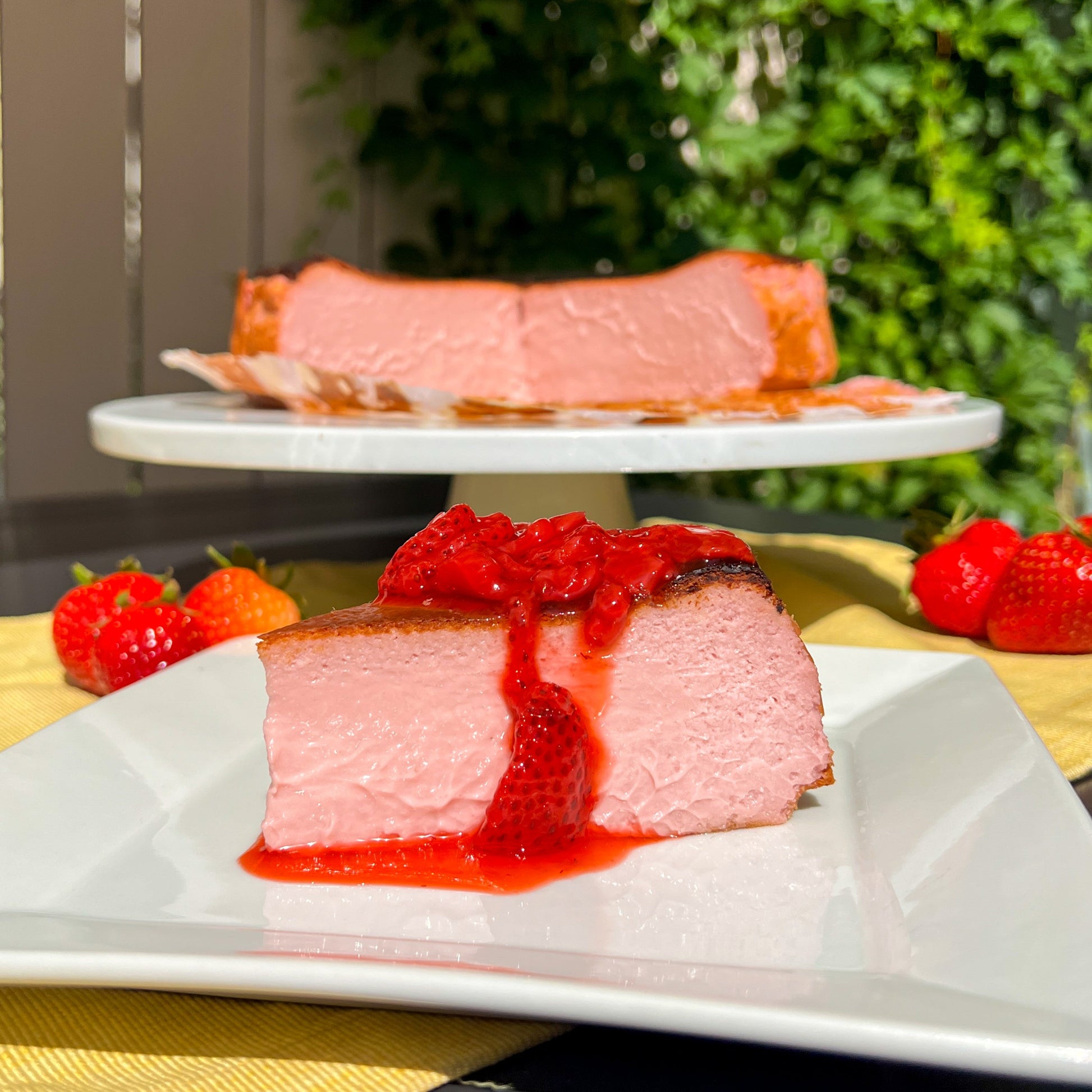 Slice of cheesecake with strawberry sauce on a white plate, with the rest of the cheesecake in the background.